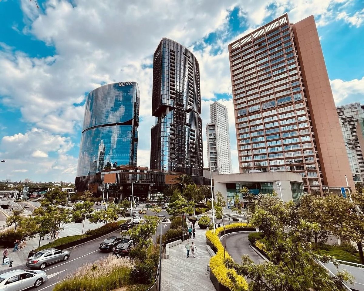 Modern urban skyline with multiple high-rise residential towers, landscaped plaza with vegetation, and clear blue sky with white clouds